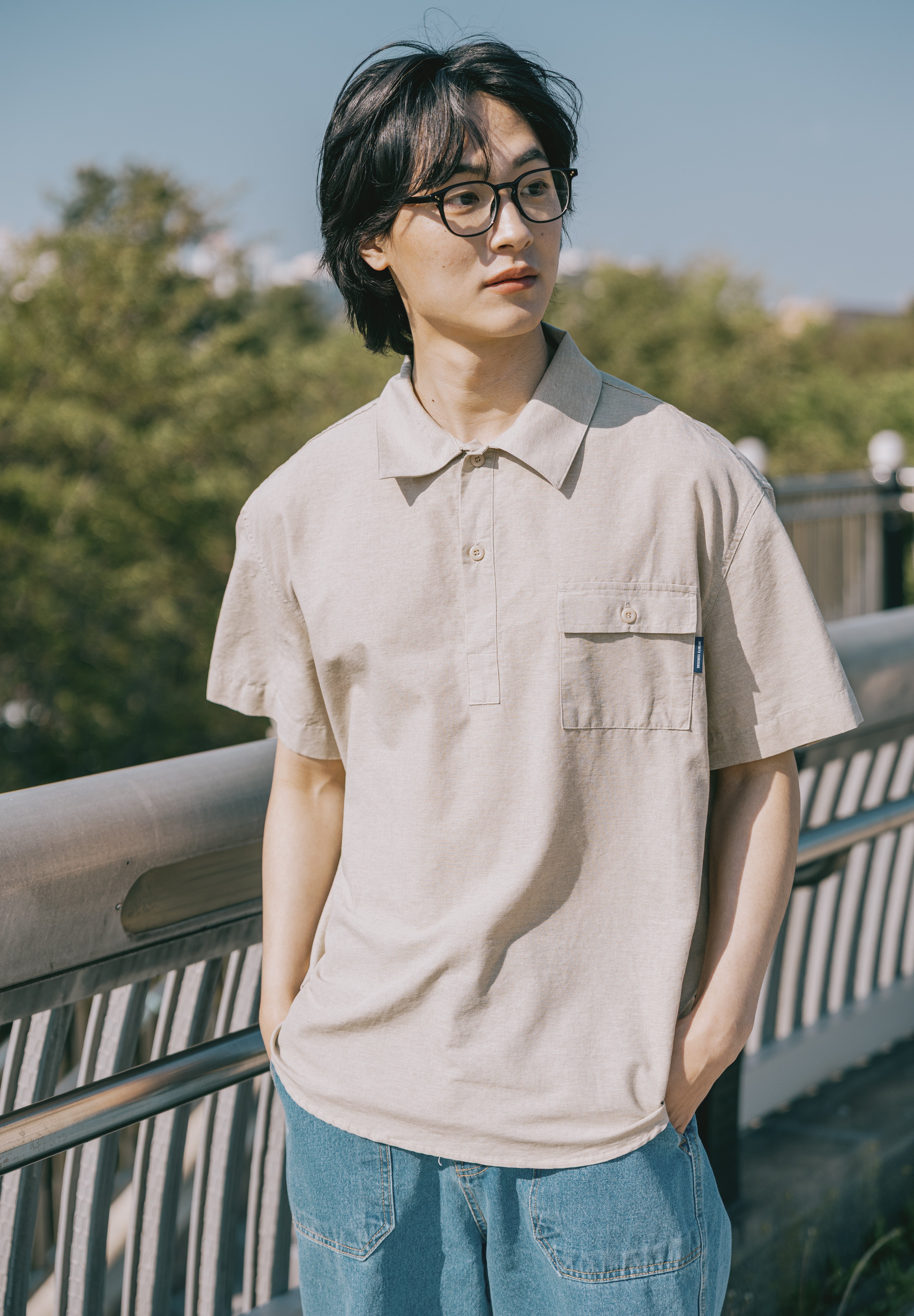 Person wearing a beige shirt and blue jeans standing outdoors with trees in the background