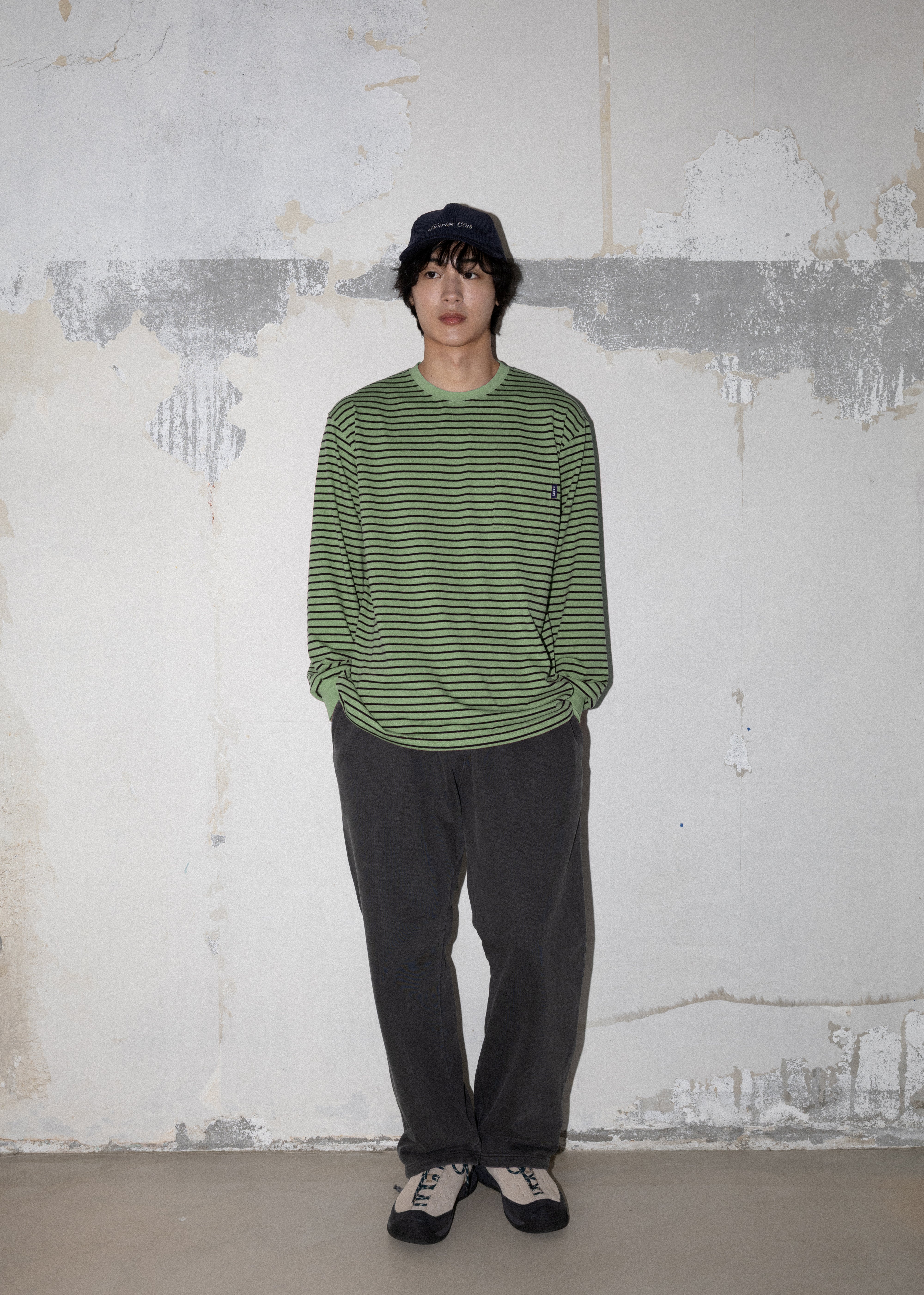 A model stands against a textured, peeling white wall wearing a green and black horizontally striped long-sleeve knit shirt, dark gray pants, and black-and-white sneakers.
